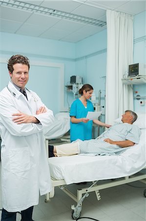 simsearch:6109-06196363,k - Smiling male doctor with his arms folded in the foreground of a room Stock Photo - Premium Royalty-Free, Code: 6109-06196364