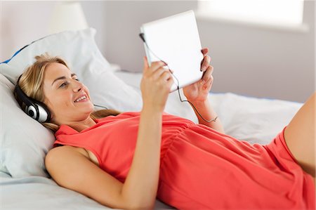 simsearch:6109-07497438,k - Happy blonde wearing dress lying on bed listening to music with tablet at home in bedroom Stock Photo - Premium Royalty-Free, Code: 6109-07497434