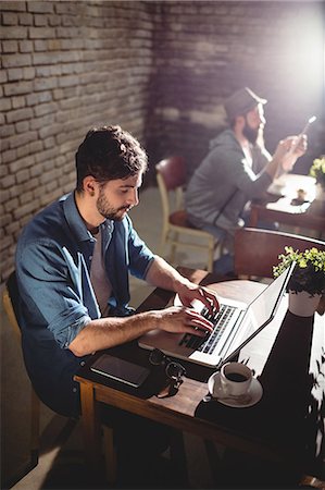 simsearch:6109-08690411,k - Handsome young man working on laptop with customer in background at cafe Stock Photo - Premium Royalty-Free, Code: 6109-08690410