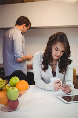 Woman using mobile phone while man working in background at kitchen Stock Photo - Premium Royalty-Free, Code: 6109-08804796