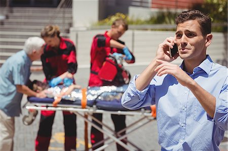 Man talking on mobile phone and paramedics examining injured boy in background Stock Photo - Premium Royalty-Free, Code: 6109-08830423