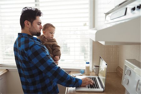 simsearch:6116-07236010,k - Father using laptop while holding his baby in kitchen at home Stock Photo - Premium Royalty-Free, Code: 6109-08944661