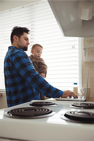 simsearch:400-05751173,k - Father using laptop while holding his baby in kitchen at home Stock Photo - Premium Royalty-Free, Code: 6109-08944662