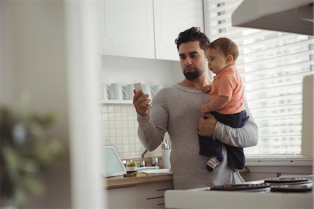simsearch:6116-07236010,k - Father using mobile phone while holding his baby in kitchen at home Stock Photo - Premium Royalty-Free, Code: 6109-08944677