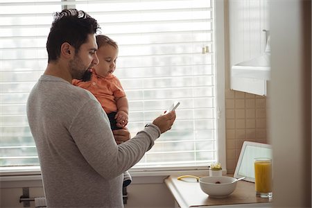 simsearch:6116-07236010,k - Father using mobile phone while holding his baby in kitchen at home Stock Photo - Premium Royalty-Free, Code: 6109-08944681