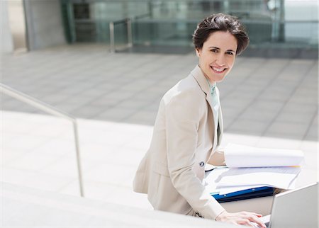 Portrait of smiling businesswoman with laptop and paperwork outside building Stock Photo - Premium Royalty-Free, Code: 6113-06497903