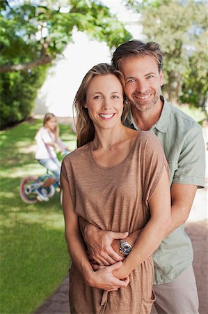 simsearch:6113-06498556,k - Portrait of smiling couple hugging in driveway with daughter on bicycle in background Stock Photo - Premium Royalty-Free, Code: 6113-06498072