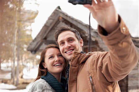 Smiling couple taking self-portrait with digital camera outside snowy cabin Stock Photo - Premium Royalty-Free, Code: 6113-06899454