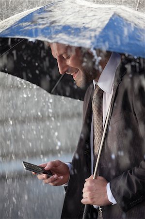 Businessman text messaging on cell phone under umbrella in rain Stock Photo - Premium Royalty-Free, Code: 6113-06899545