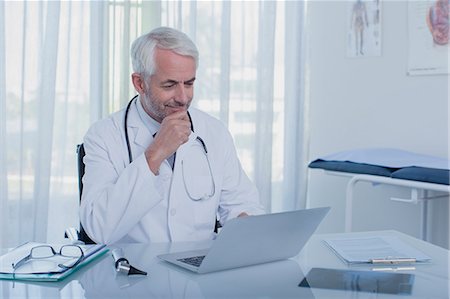 simsearch:6108-06907153,k - Smiling mature doctor sitting at desk with laptop in office Stock Photo - Premium Royalty-Free, Code: 6113-07808710