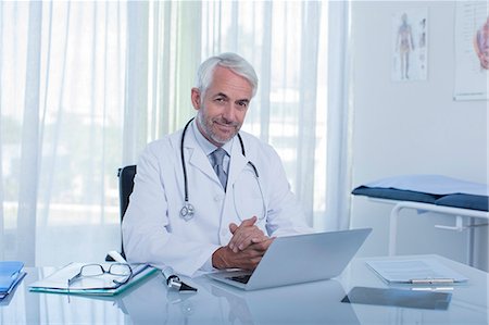 simsearch:6108-06907153,k - Portrait of smiling mature doctor sitting at desk with laptop in office Stock Photo - Premium Royalty-Free, Code: 6113-07808717