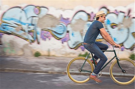Hipster man riding bicycle on road along urban graffiti wall Stock Photo - Premium Royalty-Free, Code: 6113-08171339