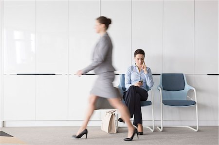 Businesswoman texting with cell phone in lobby behind walking businesswoman on the move Stock Photo - Premium Royalty-Free, Code: 6113-08171458