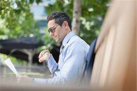 Corporate businessman eating lunch and reading paperwork on park bench Stock Photo - Premium Royalty-Free, Code: 6113-08568688