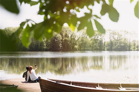 Couple with camera phone taking selfie on sunny lakeside dock next to canoe Stock Photo - Premium Royalty-Free, Code: 6113-08743425