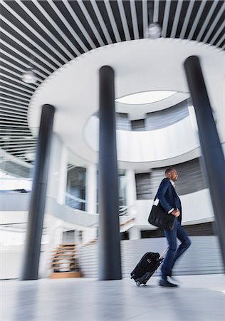 Businessman rushing, pulling suitcase in architectural, modern office lobby Stock Photo - Premium Royalty-Free, Code: 6113-09058720