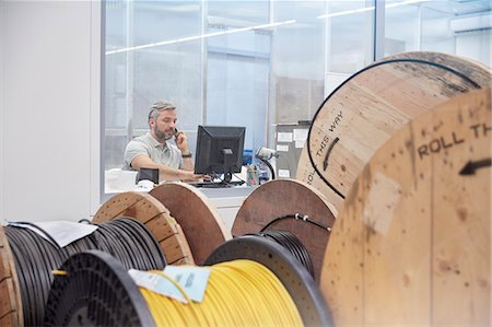 simsearch:614-08030536,k - Male supervisor working at computer and talking on ell phone behind spools in fiber optics factory Stock Photo - Premium Royalty-Free, Code: 6113-09005302