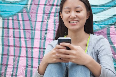 Young smiling woman sitting against a wall with graffiti and looking down and texting on her phone Stock Photo - Premium Royalty-Free, Code: 6116-07235956