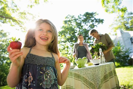 summer light - On The Farm. Children And Adults Together. A Young Girl Holding A Large Fresh Organically Produced Strawberry Fruit. Two Adults Beside A Round Table. Foto de stock - Sin royalties Premium, Código: 6118-07121831