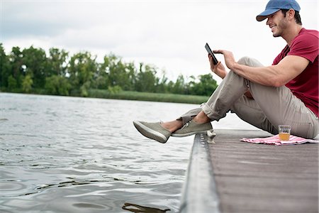 A man seated on a jetty by a lake, using a digital tablet. Stockbilder - Premium RF Lizenzfrei, Bildnummer: 6118-07351271