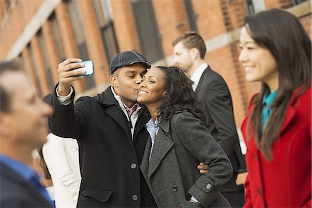 City life. A group of people on the go. A man holding out a camera phone and taking pictures of the group. Kissing a young woman. Men and women. Stock Photo - Premium Royalty-Free, Code: 6118-07353693