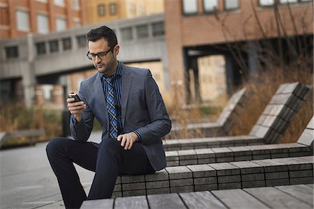 simsearch:6118-07440408,k - A man in a formal jacket and tie, sitting on a bench outside a city building, checking his phone for messages. Stock Photo - Premium Royalty-Free, Code: 6118-07353619
