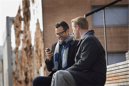 people looking phones - Two men sitting on a bench outside a large building in the city. One checking his mobile phone. Stock Photo - Premium Royalty-Free, Code: 6118-07353627