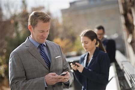 simsearch:6118-07440408,k - Three people standing on the sidewalk in the city, checking their phones. Two men and a woman. Stock Photo - Premium Royalty-Free, Code: 6118-07353633