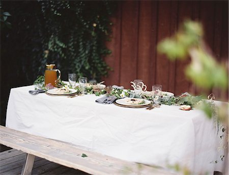 simsearch:6118-07354433,k - A table laid for a special meal. Place settings, with plates and cutlery. Glasses. A white table cloth and bench seat. Plates of food. Stock Photo - Premium Royalty-Free, Code: 6118-07354377