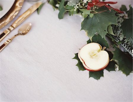 simsearch:6118-07441027,k - A table top covered with  a white cloth. A place setting with cutlery and plate. An apple cut in half, and green foliage table decoration. Photographie de stock - Premium Libres de Droits, Code: 6118-07354374