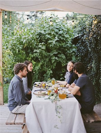 Four people seated at a table in the garden. Place settings and decorations on a white tablecloth. Two men and two women. Stock Photo - Premium Royalty-Free, Code: 6118-07354434