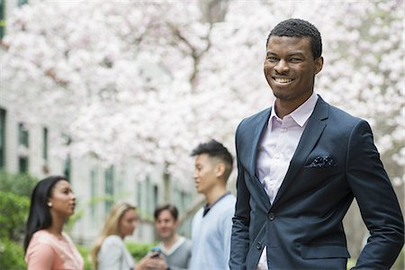 simsearch:6118-07351124,k - City life in spring. Young people outdoors in a city park. A man in a suit, smiling. Four people in the background. Using cell phones. Stock Photo - Premium Royalty-Free, Code: 6118-07354552