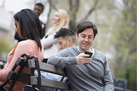 City life in spring. Young people outdoors in a city park. Sitting on a park bench. Five people, men and women, checking their phones. Stock Photo - Premium Royalty-Free, Code: 6118-07354562