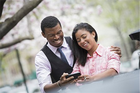 simsearch:6118-07354515,k - City life in spring. Young people outdoors in a city park. A couple side by side, with his arm around her shoulders, looking at a smart phone and smiling. Stock Photo - Premium Royalty-Free, Code: 6118-07354579