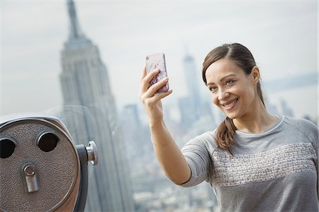 district - New York City. An observation deck overlooking the Empire State Building. A woman using her smart phone to take a photograph of herself and the view over the city. Stock Photo - Premium Royalty-Free, Code: 6118-07354513