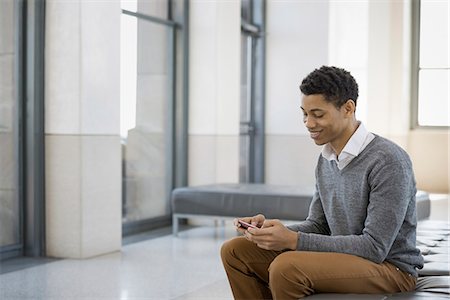 simsearch:6118-07354515,k - Urban Lifestyle. A young man sitting in a lobby, on a bench seat. Using his mobile phone. Stock Photo - Premium Royalty-Free, Code: 6118-07354520