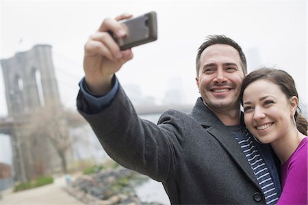 fashionable couple - New York city. The Brooklyn Bridge crossing over the East River. A couple taking a picture with a phone, a selfy of themselves. Stock Photo - Premium Royalty-Free, Code: 6118-07354645