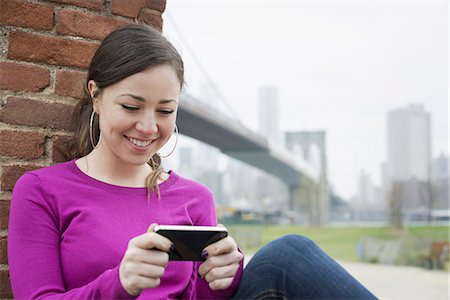 simsearch:6118-07354515,k - New York city, the Brooklyn Bridge crossing over the East River. A woman sitting leaning against a brick wall, checking her smart phone. Stock Photo - Premium Royalty-Free, Code: 6118-07354651