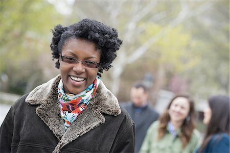 A group of people in a city park. A young woman in a coat with a large collar, smiling and looking at the camera. Stock Photo - Premium Royalty-Free, Code: 6118-07354639