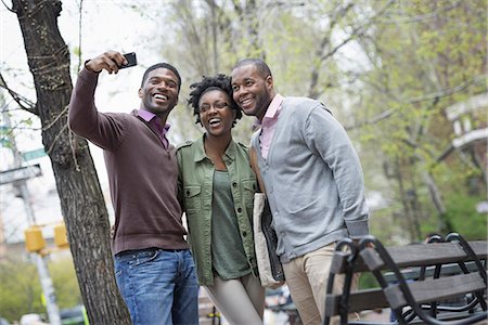 Outdoors in the city in spring. An urban lifestyle. Three people posing together and one taking a photograph of them with a smart phone. Foto de stock - Sin royalties Premium, Código: 6118-07354737