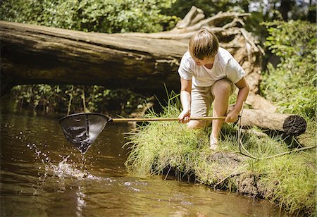 A boy kneeling by the river bank, leaning over and using a small fishing net. Stock Photo - Premium Royalty-Free, Code: 6118-07521782