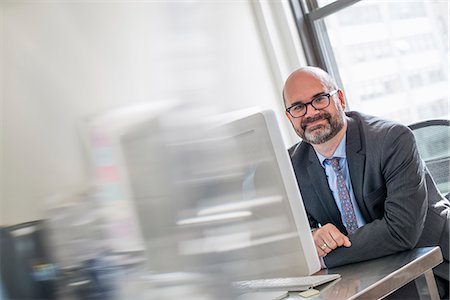 Office life. A man in a suit and tie sitting at his desk looking around his computer. Stock Photo - Premium Royalty-Free, Code: 6118-07781699