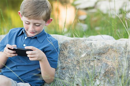 simsearch:649-03775589,k - A young boy outdoors sitting leaning against a rock, using a handheld electronic game. Stock Photo - Premium Royalty-Free, Code: 6118-07732036