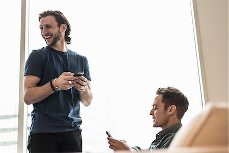 simsearch:6118-08243810,k - Two men in an office, checking their smart phones. One looking away laughing. Stock Photo - Premium Royalty-Free, Code: 6118-08066729