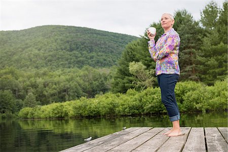 A woman standing on a lake dock holding a coffee cup. Stock Photo - Premium Royalty-Free, Code: 6118-08243933