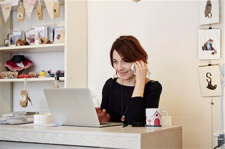 simsearch:6118-08202525,k - A woman sitting at a desk in a gift shop using a laptop and making a call. Stock Photo - Premium Royalty-Free, Code: 6118-08202518