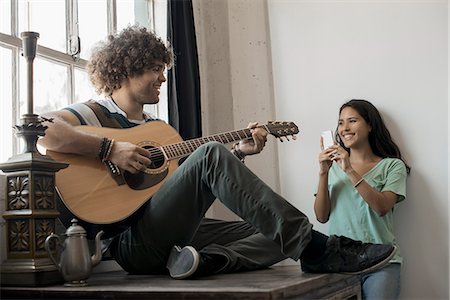 Loft living. A young man playing guitar to an appreciative audience, a young woman holding a smart phone and taking a photograph. Stock Photo - Premium Royalty-Free, Code: 6118-08394001