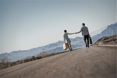 future of the desert - A young couple, man and woman walking hand in hand on a tarmac road in the desert carrying cases. Stock Photo - Premium Royalty-Free, Code: 6118-08394226