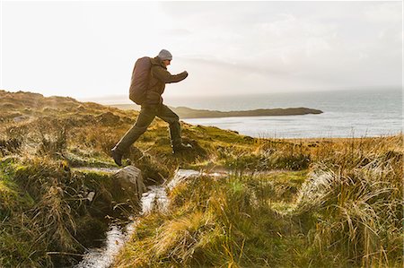 simsearch:6118-08399701,k - A man with a rucksack and winter clothing leaping across a small stream in an open exposed landscape. Stock Photo - Premium Royalty-Free, Code: 6118-08399672