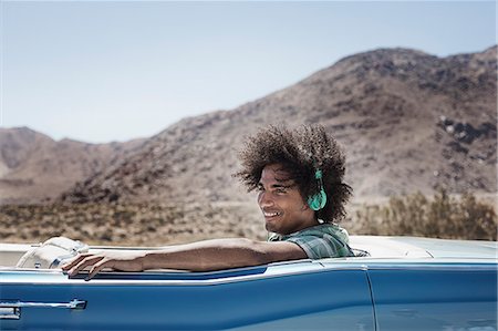 A young man with music headphones sitting in the back of a pale blue convertible on the open road in the mountains. Stock Photo - Premium Royalty-Free, Code: 6118-08488179